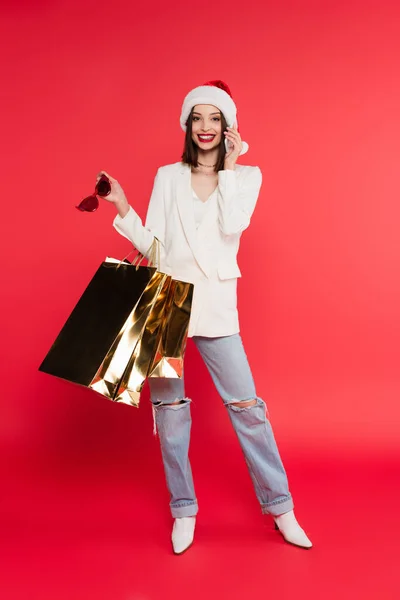 Mujer alegre en sombrero de santa celebración de bolsas de compras y gafas de sol mientras habla en el teléfono inteligente sobre fondo rojo - foto de stock