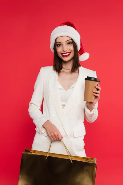 Stylish woman in santa hat holding shopping bag and coffee to go isolated on red — Stock Photo