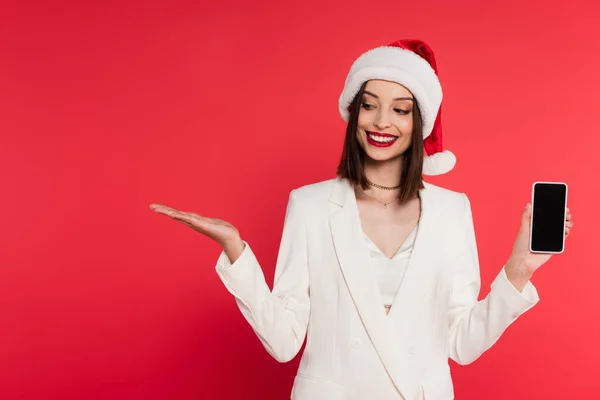 Stylische Frau mit Weihnachtsmannmütze hält Handy mit leerem Bildschirm und zeigt mit der Hand isoliert auf rot — Stockfoto