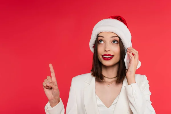 Smiling woman with red lips and santa hat talking on smartphone and pointing with finger isolated on red — Stock Photo