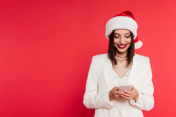 Mulher positiva em chapéu de santa e jaqueta branca usando telefone celular isolado em vermelho — Fotografia de Stock