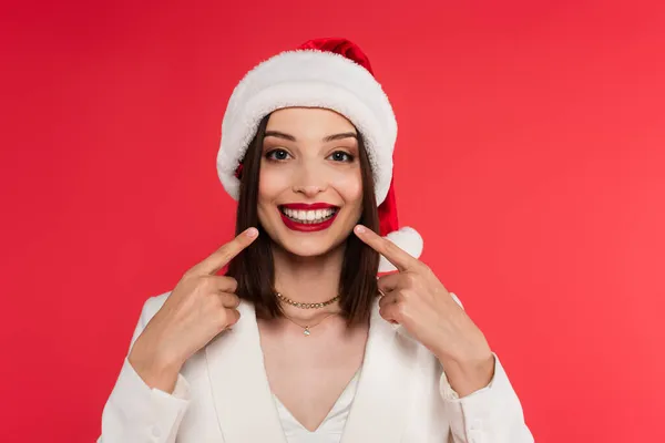 Усміхнена жінка в капелюсі Санти вказує на зуби ізольовані на червоному — стокове фото