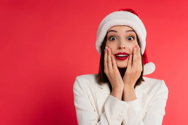 Donna stupita con labbra rosse e cappello di Babbo Natale guardando la fotocamera isolata sul rosso — Foto stock