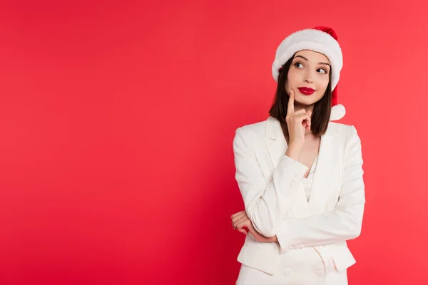 Pensive woman in santa hat and white jacket looking away isolated on red — Stock Photo