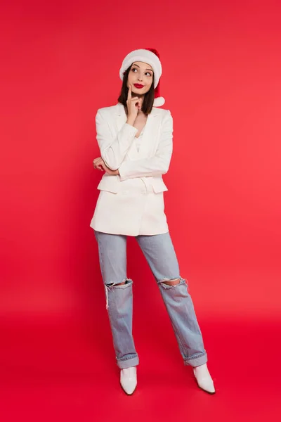 Full length of pensive woman in santa hat and white jacket on red background — Stock Photo