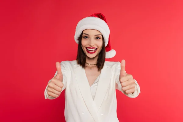 Mujer positiva con labios rojos y sombrero de santa mostrando los pulgares hacia arriba aislado en rojo - foto de stock