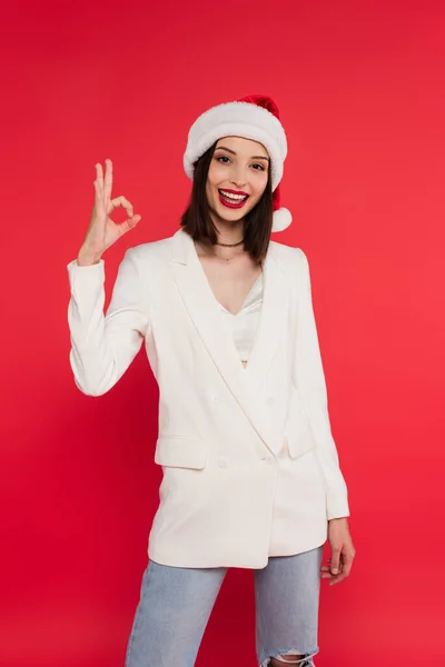 Stylish woman in santa hat showing okay gesture isolated on red — Stock Photo