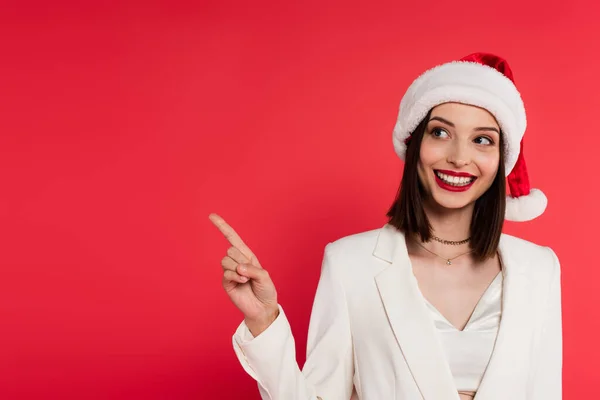 Cheerful woman in santa hat pointing with finger isolated on red — Stock Photo