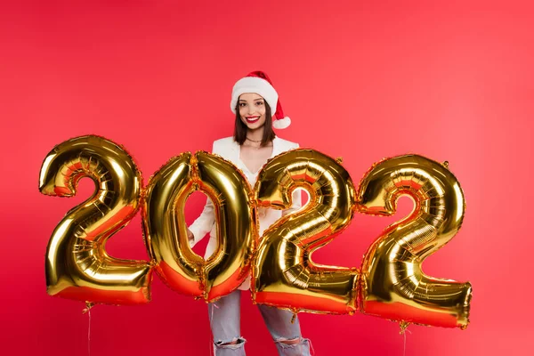 Mujer bonita en sombrero de santa de pie cerca de globos en forma de 2022 números aislados en rojo - foto de stock