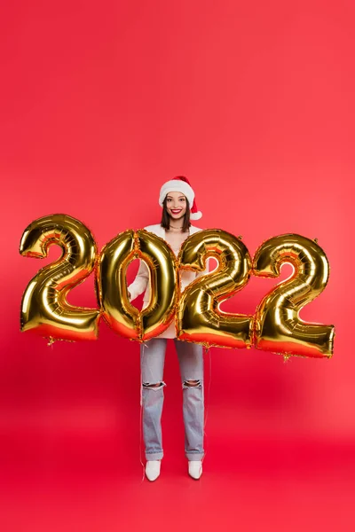 Mujer joven en sombrero de santa de pie cerca de globos en forma de 2022 números sobre fondo rojo - foto de stock