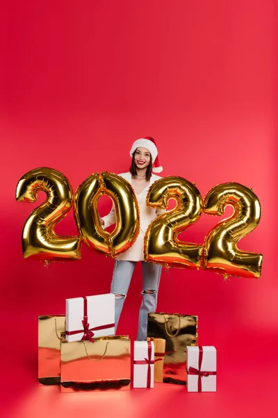 Mujer feliz en sombrero de santa de pie cerca de globos en forma de 2022 números, regalos y bolsas de compras sobre fondo rojo - foto de stock