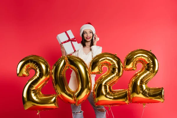 Mulher sorridente em santa chapéu falando no smartphone e segurando presente perto de balões em forma de 2022 números isolados em vermelho — Fotografia de Stock