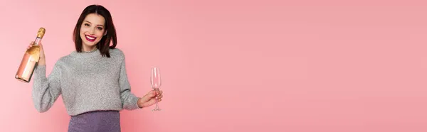 Positive woman holding bottle of champagne and glass isolated on pink, banner — Stock Photo