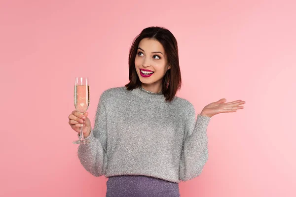 Donna sorridente in maglione che tiene il bicchiere con lo champagne e punta con mano isolata sul rosa — Foto stock