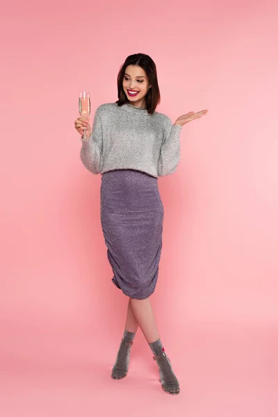 Positive Frau in Pullover und Rock hält ein Glas Champagner in der Hand und zeigt mit der Hand auf rosa Hintergrund — Stockfoto