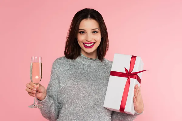Hübsche Frau mit Champagner und Geschenk blickt isoliert in die Kamera auf rosa — Stockfoto