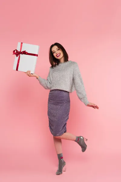 Positive woman in skirt and sweater holding present on pink background — Stock Photo