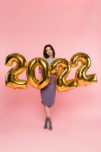 Mujer elegante sonriendo cerca de globos en forma de números 2022 sobre fondo rosa - foto de stock