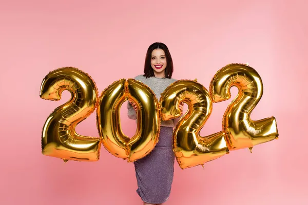 Glückliche, stylische Frau, die neben Luftballons in Form von Zahlen von 2022 steht, isoliert auf rosa — Stockfoto