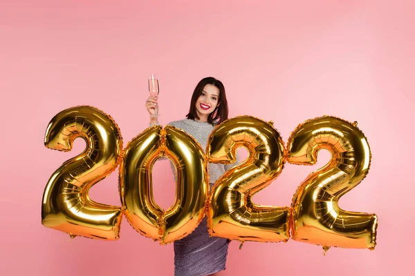 Усміхнена жінка тримає келих шампанського біля кульок у формі 2022 року ізольовано на рожевому — стокове фото