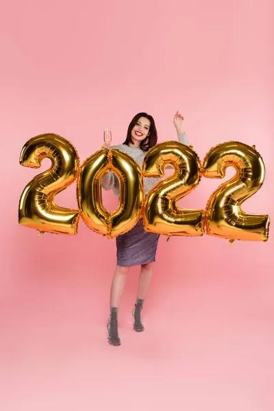 Mulher bonita e elegante segurando champanhe perto de balões em forma de 2022 no fundo rosa — Fotografia de Stock