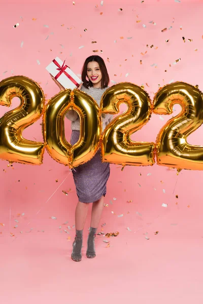 Smiling woman holding present near balloons in shape of 2022 and confetti on pink background — Stock Photo