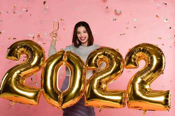 Cheerful woman holding champagne near balloons in shape of 2022 and confetti on pink background — Stock Photo