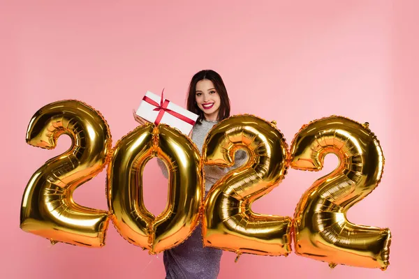 Donna sorridente in possesso di scatola regalo vicino palloncini in forma di 2022 numeri isolati su rosa — Foto stock