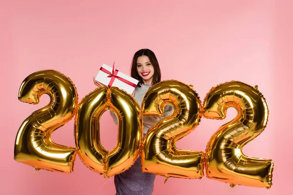 Smiling woman holding gift box near balloons in shape of 2022 numbers isolated on pink — Stock Photo