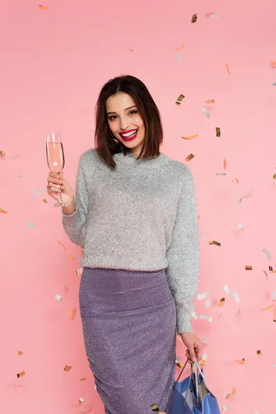 Trendy woman with champagne and shopping bags standing under confetti on pink background — Stock Photo