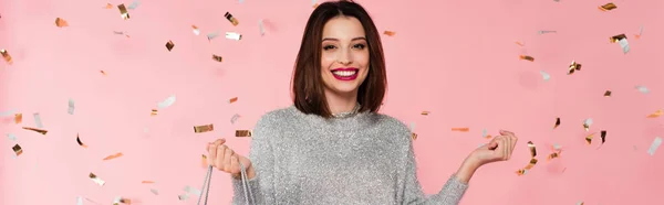 Positive woman in sweater holding shopping bag under confetti on pink background, banner — Stock Photo