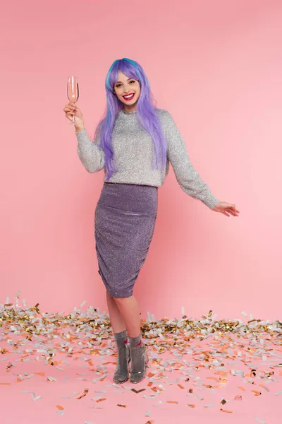 Happy stylish woman with dyed hair holding champagne near confetti on pink background — Stock Photo