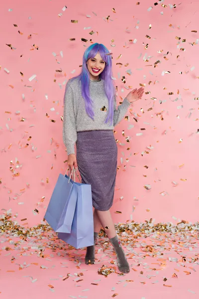 Mulher positiva com cabelos tingidos segurando sacos de compras e acenando a mão sob confete no fundo rosa — Fotografia de Stock