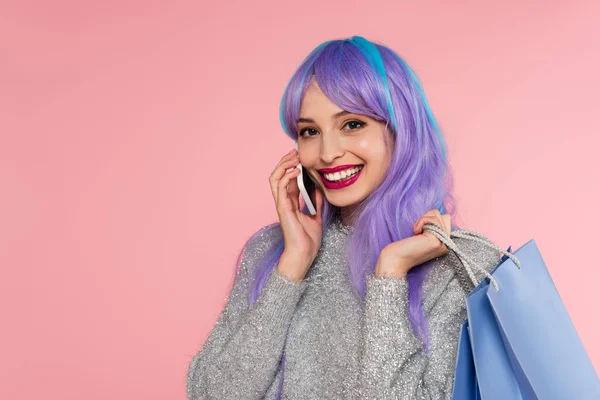 Mujer elegante con el pelo teñido hablando en el teléfono inteligente y la celebración de bolsas de compras aisladas en rosa - foto de stock