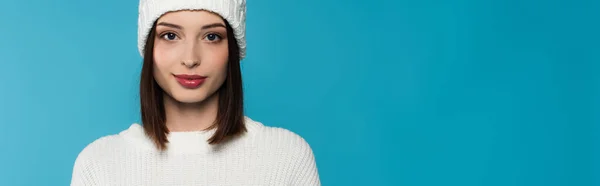 Brunette woman in knitted hat and sweater looking at camera isolated on blue, banner — Stock Photo