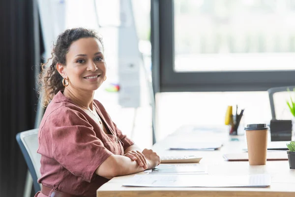 African american businesswoman smiling at camera near papers and coffee to go in office — Stock Photo
