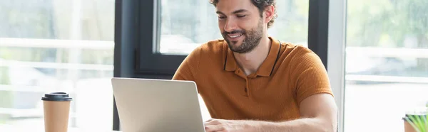 Positiver Geschäftsmann arbeitet am Laptop in der Nähe von Coffee to go im Büro, Banner — Stockfoto