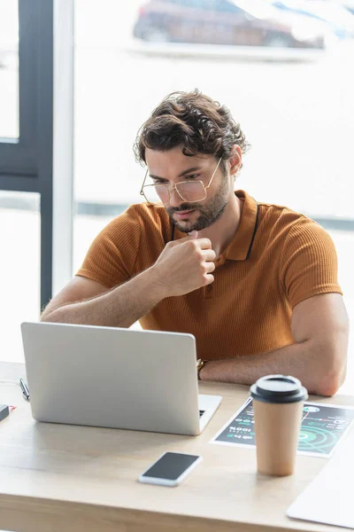 Empresário em óculos usando laptop perto de celular e bebida takeaway no escritório — Fotografia de Stock