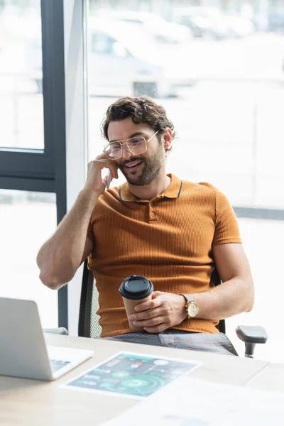 Empresário alegre em óculos falando no smartphone e segurando café para ir perto do laptop no escritório — Fotografia de Stock