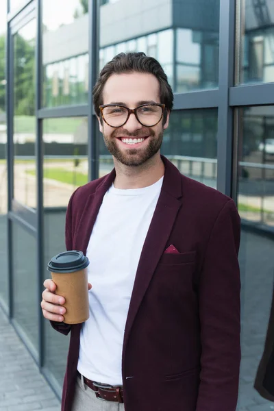 Uomo d'affari sorridente con caffè per andare a sorridere alla macchina fotografica vicino a costruire all'aperto — Foto stock