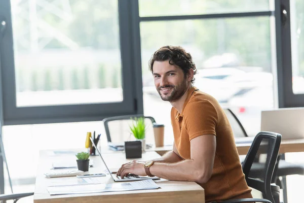 Hombre de negocios positivo mirando a la cámara mientras usa el ordenador portátil cerca de los papeles en la oficina - foto de stock