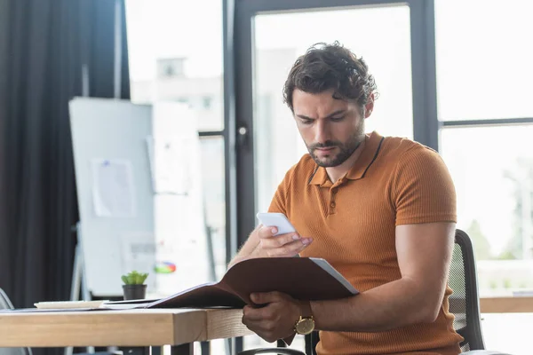 Businessman using smartphone and holding paper folder near working table in office — Stock Photo