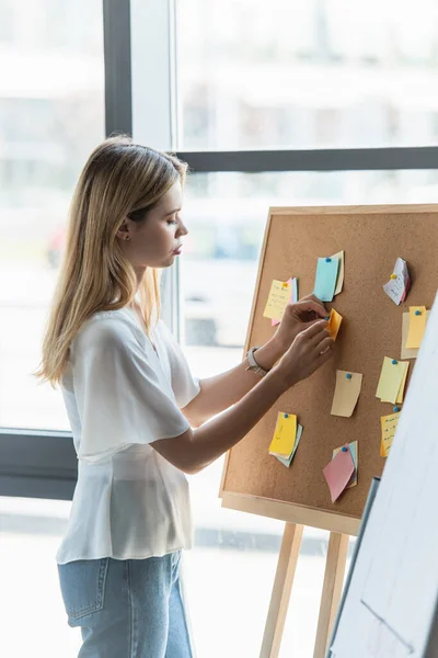 Side view of young businesswoman pinning sticky notes on board in office — Stock Photo