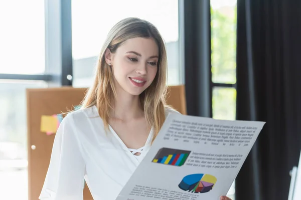 Empresária feliz olhando para papel desfocado com gráficos no escritório — Fotografia de Stock