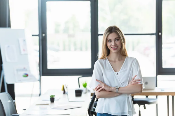 Jeune femme d'affaires avec les bras croisés souriant à la caméra dans le bureau — Photo de stock