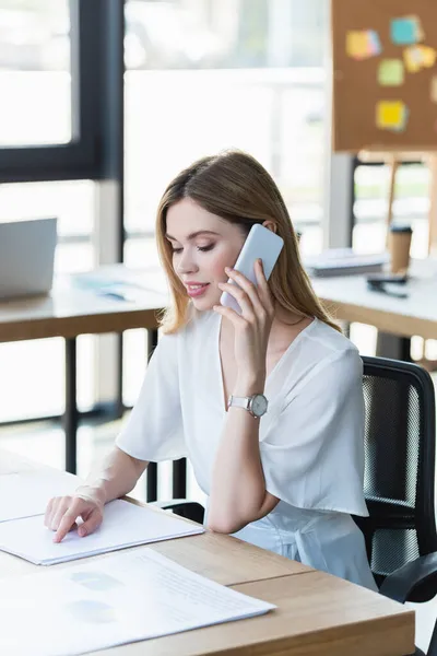 Empresária positiva falando no smartphone e apontando para o documento na mesa de escritório — Stock Photo