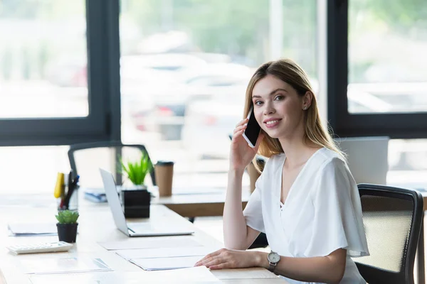 Junge Geschäftsfrau lächelt, während sie neben Dokumenten auf dem Tisch mit dem Handy telefoniert — Stockfoto