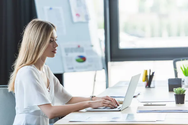 Joven mujer de negocios utilizando el ordenador portátil cerca de documentos y teléfonos inteligentes en la oficina — Stock Photo