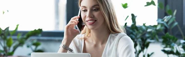 Feliz mujer de negocios hablando en el teléfono inteligente cerca de la computadora portátil en la oficina, bandera — Stock Photo