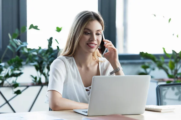 Empresária alegre falando no celular e usando laptop no escritório — Fotografia de Stock